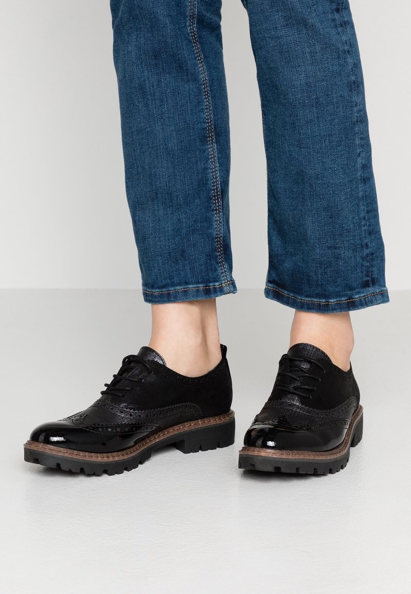 Henkilö, jolla on päällä lyhyet siniset farkut ja mustat nauhalliset brogue-kengät, joissa on paksut pohjat, vaalealla lattialla.