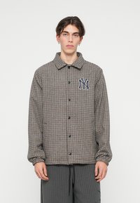 Jachetă în carouri maro și crem, cu guler, închidere cu nasturi și un logo NY bleu pe pieptul stâng. Mâneci lungi cu manșete elastice.