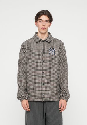 Chaqueta a cuadros marrón y crema con cuello, cierre de botones y un logo de NY en azul marino en el pecho izquierdo. Mangas largas con puños elásticos.