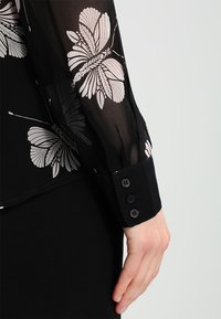 Blusa nera trasparente con stampe di farfalle bianche, caratterizzata da polsini neri solidi con tre bottoni, che mettono in evidenza una delicata texture e un design floreale.