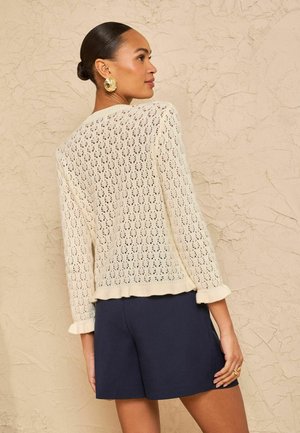 Femeie cu părul prins în coc poartă cercei mari de aur, un pulover tricotat crem cu model asemănător dantelei și pantaloni scurți bleumarin, stând în picioare lângă un perete texturat.