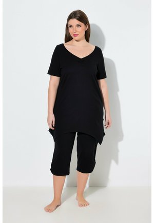 Donna in piedi a piedi nudi che indossa una tunica nera a maniche corte con orlo asimmetrico e pantaloni neri corti davanti a uno sfondo bianco.