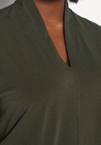 Olivi vihreä pusero, jossa on v-aukkoinen kaula-aukko, sileä rakenne ja hienovarainen laskostus. Ei näkyviä kuvioita tai varusteita.