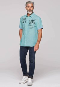 Helblau groen T-shirt met korte mouwen en prints, knoopsluiting aan de voorkant, en contrasterende blauwe manchetten. Gecombineerd met donkere spijkerbroeken en witte sneakers.