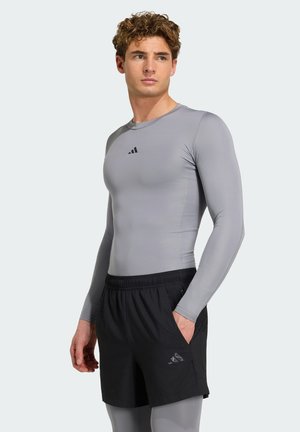 Jeune homme aux cheveux bouclés portant un t-shirt gris cintré à manches longues, un short de sport noir et des leggings gris, debout avec la main dans la poche.