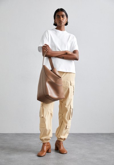 Sac en cuir marron décontracté avec un dessus incurvé et une bandoulière, associé à un t-shirt blanc et un pantalon cargo beige.