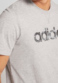 Grå bomullströja med en framträdande svart och grå mönstrad "adidas"-logotyp över bröstet. Kort ärm, rund halsdesign.