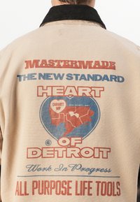 Bežs kanvas žakets ar lielu grafiku aizmugurē, kurā ir sarkans un zils teksts, kurā ir uzraksts "Mastermade Heart of Detroit" un kartes kontūra.