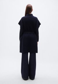 Μπλε σκούρο πλεκτό φαρδύ γιλέκο με γιακά κασκόλ, ζώνη στη μέση και φαρδιά μανίκια, σετάρισμένο με τζιν παντελόνι με φαρδιά μπότα στο ίδιο χρώμα.