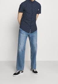 Tmavě modrá košile s krátkými rukávy na knoflíky s bílými a červenými puntíky, ve které jsou světlé modré džíny s širokými nohavicemi a černé tenisky s bílými detaily.