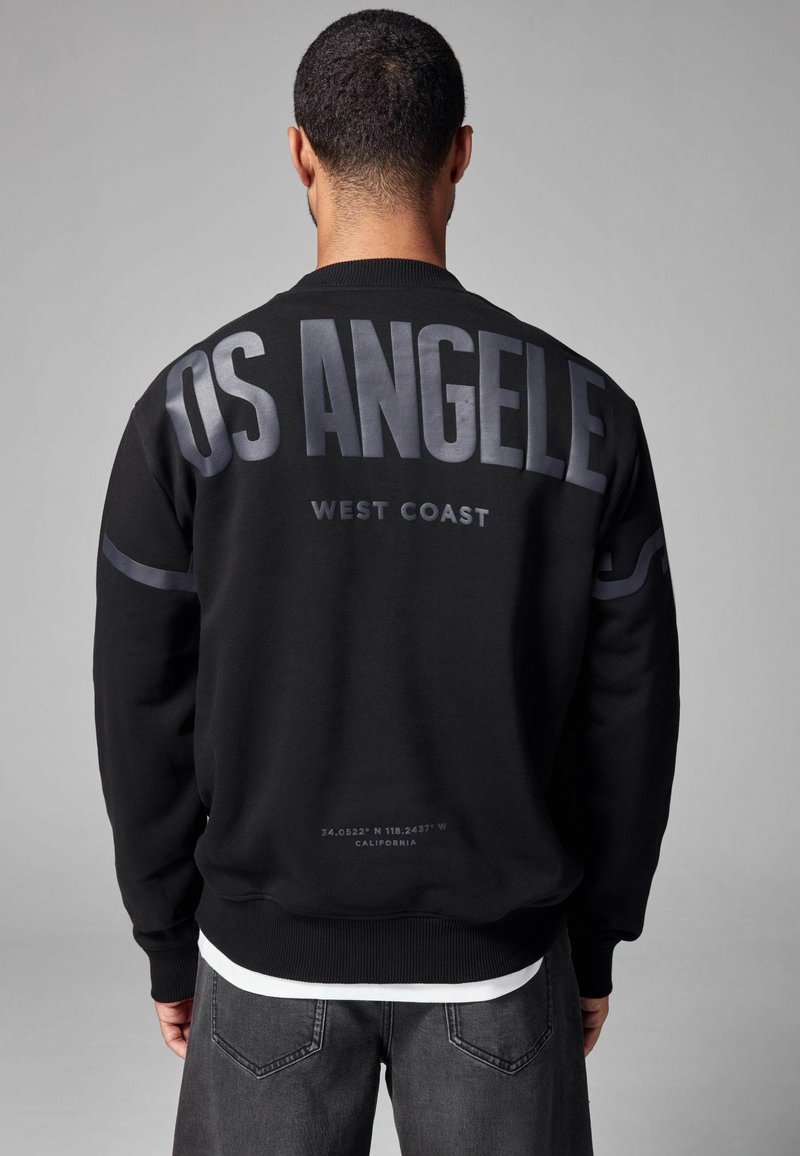 Čierna mikina so širokým potlačou "LOS ANGELES" na zadnej strane, s okrúhlym výstrihom, patentnými manžetami a jemnými textúrami.
