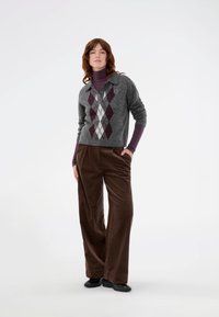 Grauer Pullover mit Rautenmuster in Burgunderrot und Weiß, kombiniert mit braunen Cordhosen im weiten Schnitt und schwarzen Schuhen. Das Model steht mit den Händen in den Taschen.