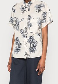Chemise à manches courtes en tissu beige clair, avec des motifs floraux et de feuilles noirs, boutonnière sur le devant et poche poitrine unique.