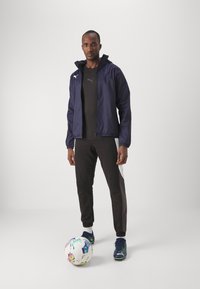 Marine windbreaker med hætte, matchende sort træningsshirt, brune joggers med en hvid sidepanel og blå fodboldskon; farverig fodbold.