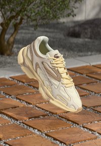 Baskets Lacoste en crème et jaune avec un design superposé, dessus léger et accents texturés, flottant au-dessus d'une surface carrelée.
