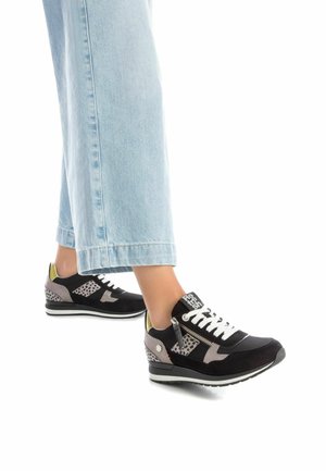 Zapatillas negras y grises con materiales de ante y malla, detalles perforados, cordones blancos y un detalle verde en el talón. Combinadas con jeans de color azul claro.