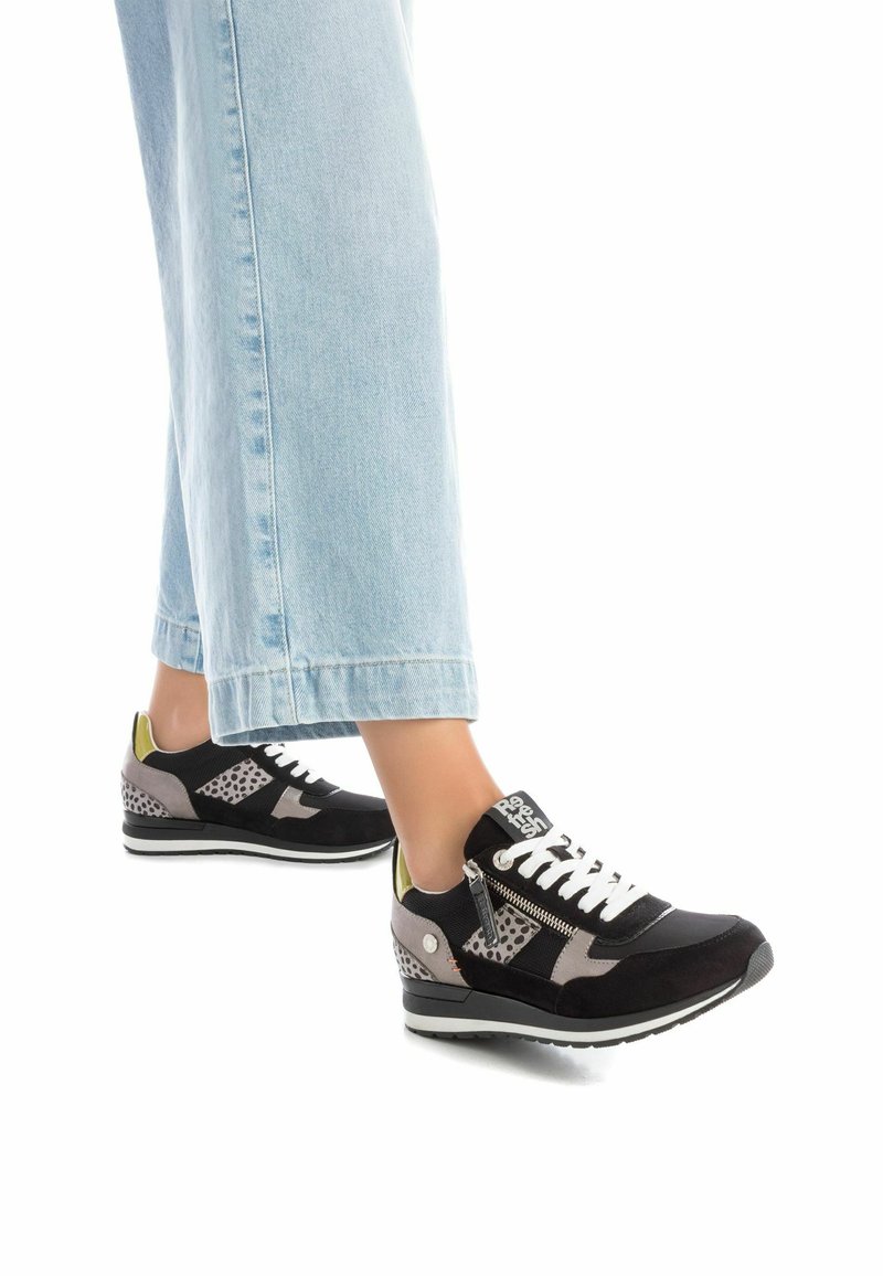 Zapatillas negras y grises con materiales de ante y malla, detalles perforados, cordones blancos y un detalle verde en el talón. Combinadas con jeans de color azul claro.