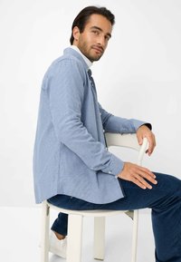 Camicia a maniche lunghe di un blu chiaro con motivo, con bottoni davanti e colletto bianco a contrasto. Indossata sopra pantaloni blu navy, seduto su una sedia bianca.