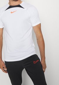 Biała koszulka sportowa Nike z czarnymi panelami na ramionach i pomarańczowym logo. Czarne spodnie z małym pomarańczowym logo na udzie.