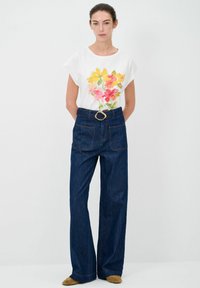 T-shirt bianco a maniche corte con design floreale colorato. Pantaloni in denim scuro a vita alta e gamba larga con tasche e fibbia dorata.