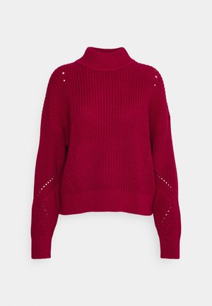 Pull en grosse maille rouge avec col montant, manches longues et petits détails ajourés aux épaules et aux manches.