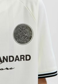 Emblema circular en blanco y negro que muestra dos figuras, una de pie y otra de rodillas, sobre una camiseta blanca con puños de mangas a rayas negras.