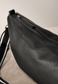 Sac bandoulière en cuir noir texturé avec fermeture éclair argentée et une sangle d'épaule réglable rayée noir et blanc sur une surface claire.