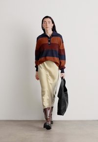 Pull en tricot avec des rayures en rouille et bleu marine, design à col, associé à une jupe beige en satin, des bottes marron montantes, et un sac tote noir.