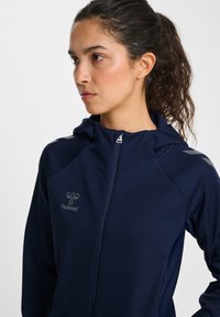 Marinblå zip-up hoodie med hög krage och en logotyp på vänster bröst. Har ljusgrå detaljer på axlarna och en len, mjuk textur.