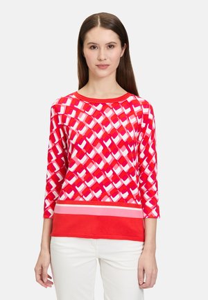 Haut à motif rouge avec des formes géométriques en rose et blanc, doté d'une bande rouge unie à l'ourlet et de manches trois-quarts. Texture de tissu douce.