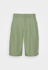 Shorts verts mi-longs avec plis, passants de ceinture, fermeture à bouton, poches latérales et petit swoosh blanc de Nike sur l'ourlet de la jambe gauche.