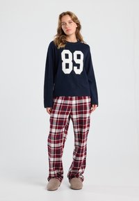Vrouw staat in een gezellige outfit met een marineblauwe trui met het nummer 89, rood-witte geruite broek en beige sloffen op een witte achtergrond.