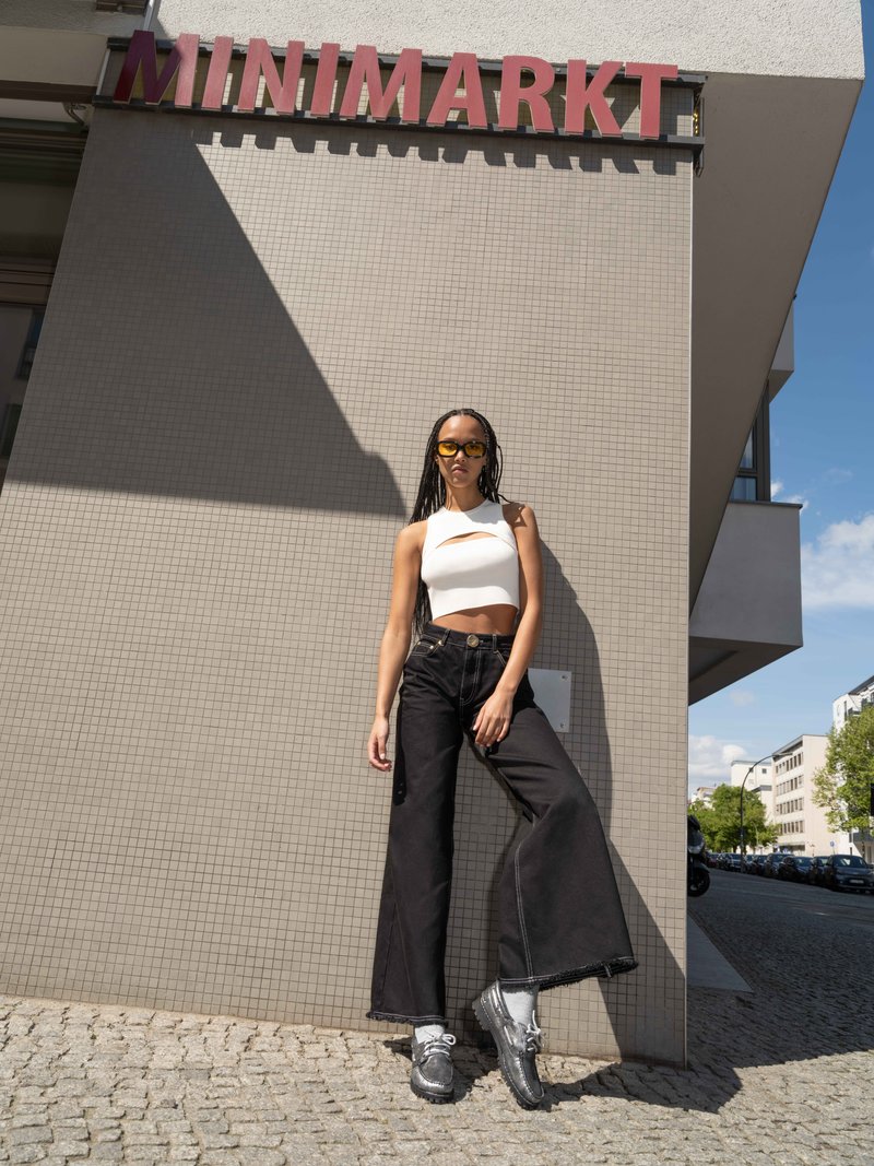 Jeans neri a gamba larga con orlo grezzo, top bianco corto e scarpe grigie a strati. Occhiali da sole gialli completano l'outfit casual davanti a una parete texturizzata.