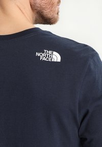 Tmavě modré bavlněné tričko s kulatým výstřihem, s bílým logem na horní části zad. Hladký povrch s krátkými rukávy a bez vzorů.