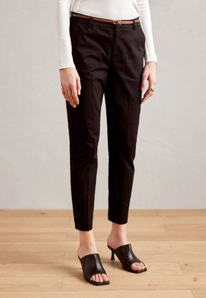 Femme portant un pantalon noir slim, une ceinture marron, un haut blanc à manches longues et des mules à talons ouvertes noires, debout sur un sol en bois.