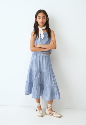 Meisje met lang donker haar staat met armen over elkaar, draagt een mouwloos blauw topje, bijpassende gelaagde rok, witte nekdoek en veterespadrilles.