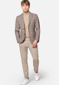 Brązowa marynarka w kratę z dwoma guzikami, beżowy golf, jasnobeżowe spodnie i białe sneakersy. Gładki materiał i dopasowany krój.