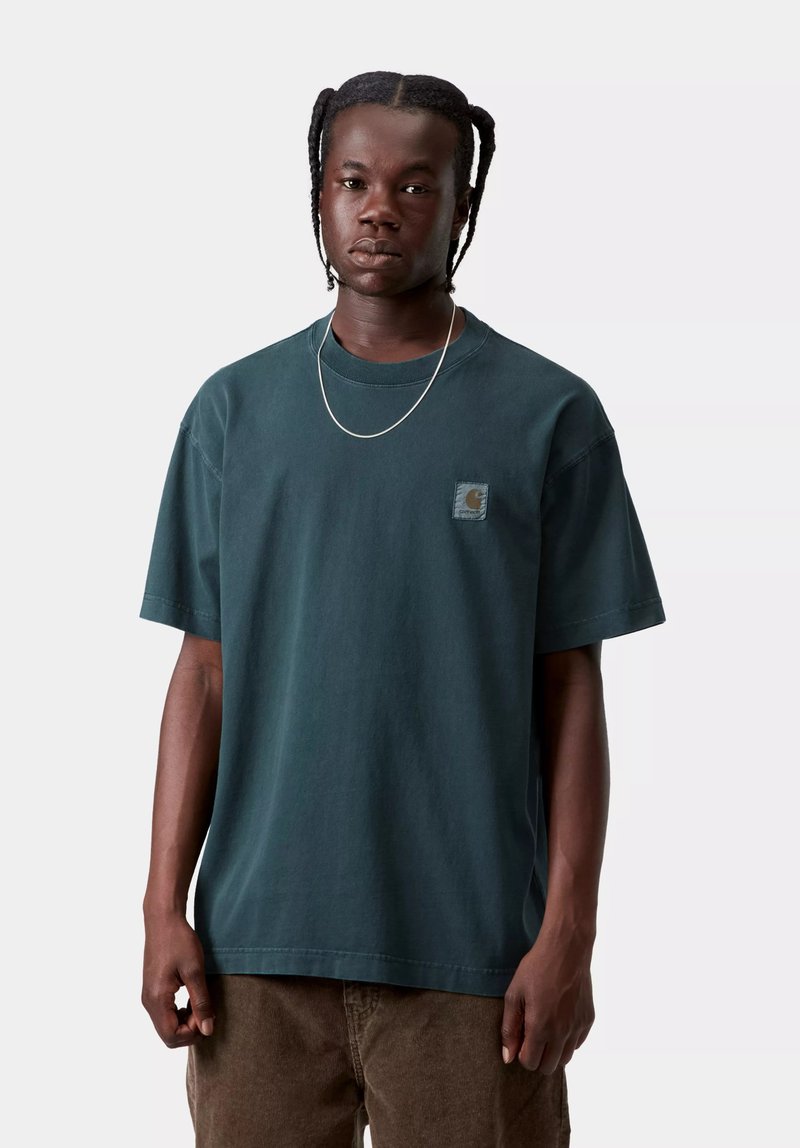 Jeune homme aux cheveux tressés portant un t-shirt Carhartt vert foncé, un collier chaîne argenté et un pantalon marron sur un fond uni.
