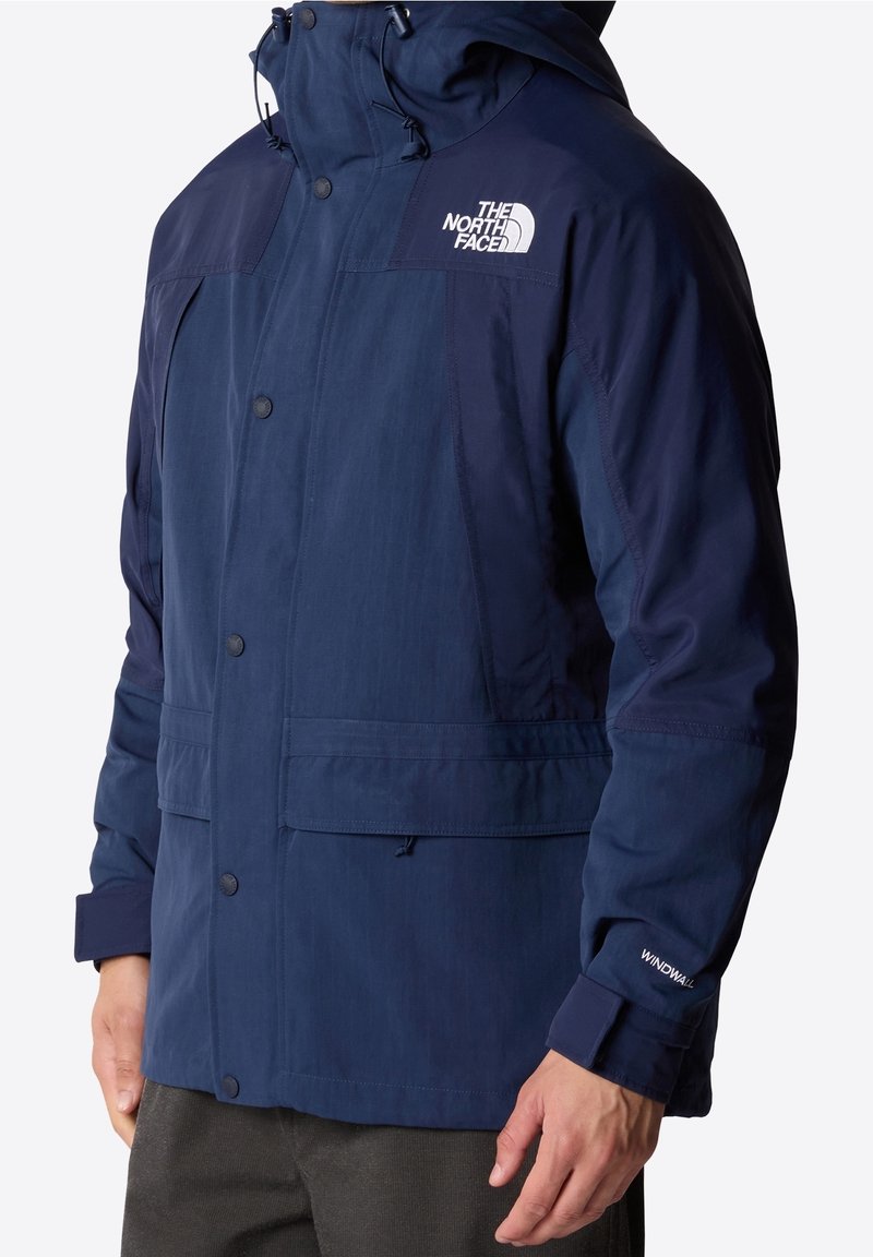 Personne portant une veste The North Face bleu foncé avec capuche, logo visible sur la poitrine et texte WindWall sur la manche, mains sur les côtés.