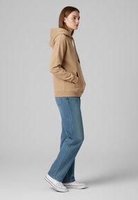 Felpa con cappuccio color khaki con tasca frontale, abbinata a jeans azzurri chiari e sneakers bianche. Il tessuto sembra morbido e ha una vestibilità comoda.