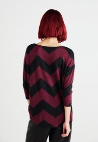 Bordeaux-rotes und schwarzes Pullover im Chevron-Muster mit weitem Halsausschnitt und 3/4-Ärmeln. Weicher Stoff, lockere Passform und abgerundeter Saum.