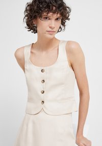 Beige ærmeløs vest med firkantet udskæring, fem brune knapper foran og sidelommer. Glat tekstur og skræddersyet pasform.