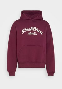 Bordeauxfärgad huvtröja med framficka och vit kursiv text "WnstBhvr Studio" på bröstet, visad mot en enkel ljus bakgrund.