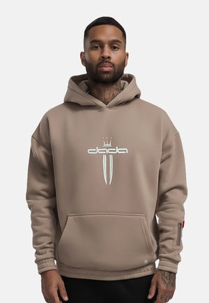 Man met baard en tatoeages draagt een beige hoodie met wit Dada-logo en voorzak, staand tegen een effen achtergrond.