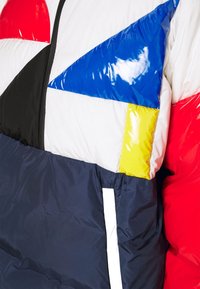 Puffy jacket with a geometric pattern featuring red, blue, yellow, white, black, and navy panels. Glossy finish, textured fabric, and pocket detail.