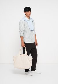 Bolso de lona en beige claro, con asas dobles, textura suave y un diseño amplio. Modelo vistiendo una camisa a rayas, look casual.
