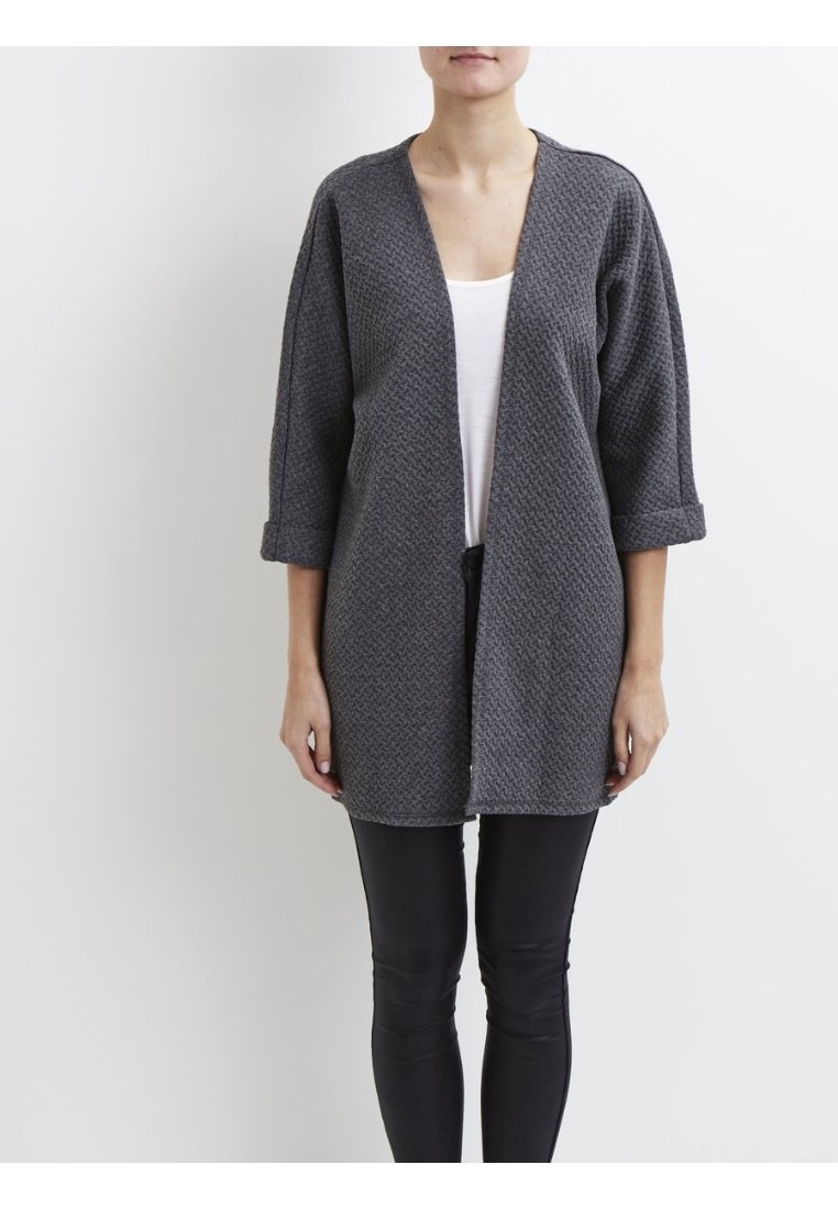 Personne portant un cardigan gris texturé à ouverture avant avec manches 3/4 retroussées, un haut blanc et un pantalon noir ajusté sur un fond blanc uni.