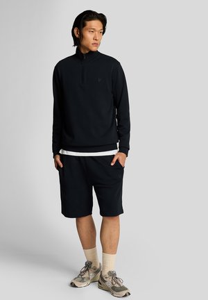 Sweat-shirt marine à fermeture éclair quart avec des poignets côtelés, associé à un short marine à hauteur des genoux. Chaussettes de couleur claire et chaussures de sport à motifs multiples.