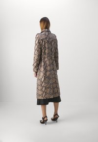 Trench-coat long à motif serpent dans des tons de beige et marron ; doté d'une ceinture à la taille, d'un col et de boutons ; porté avec des talons noirs.