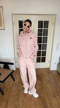 Kvinde i lyserød jakke og bukser med hvide sneakers og hvide solbriller, stående indendørs på trægulv med hænderne i lommerne.